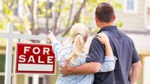 A house with a "For Sale" sign in front of it, surrounded by other houses